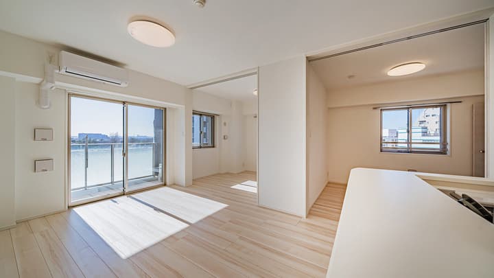 View from the kitchen area across the light wood-style floor, showing the main living area with a balcony door and an opening to an adjacent room with a window.