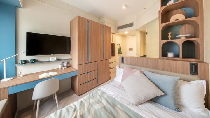 View of a compact bedroom featuring a double bed with light blue and pink pillows. The room has extensive, integrated wood-look storage and cabinetry, a small desk area with a mounted TV, and a decorative built-in shelving unit next to the bed.