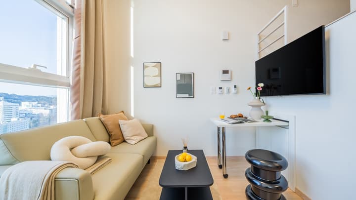 Small living area featuring a pale green couch, a black coffee table with lemons, a wall-mounted TV, and a large window with a city view.