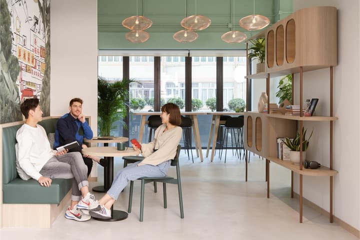 Bright, modern communal lounge area with a long wooden table, green accent wall, and residents sitting and talking in a stylish coliving space.
