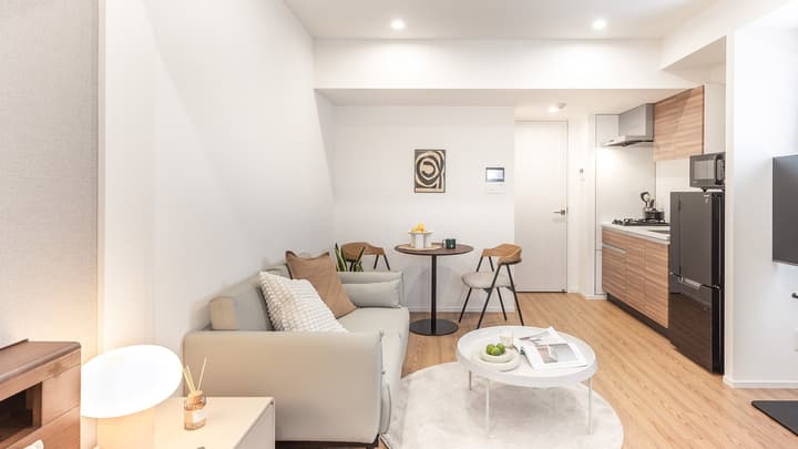 Wide view of the open-concept apartment, showing the living area with a sofa, small round dining table, and a partial view of the modern kitchenette with light wood cabinets.
