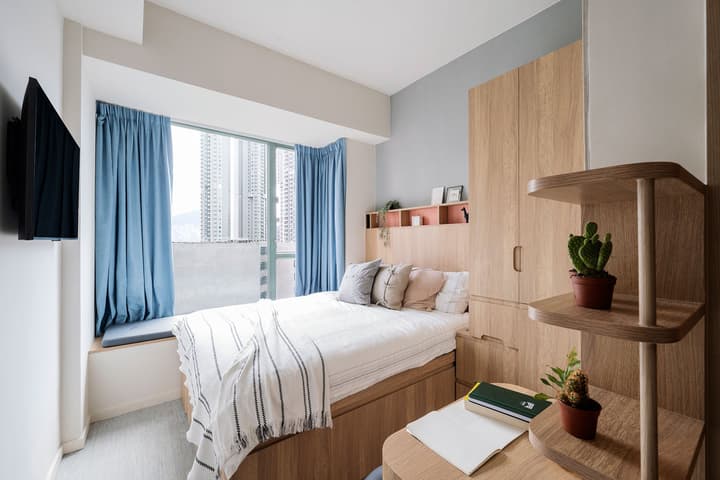 Interior view of a queen-sized bed next to a large window with light blue curtains and a cushioned window seat. The room has light wood-grain cabinetry, including a tall wardrobe and a low headboard shelf. A wall-mounted TV is visible on the far left.