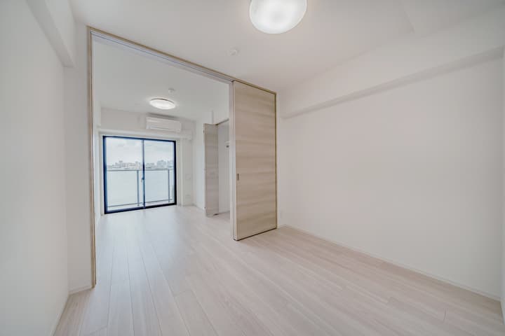 Living and dining room with light wood floors and white walls, featuring large sliding pocket doors that separate the main room from the bedroom with a balcony.