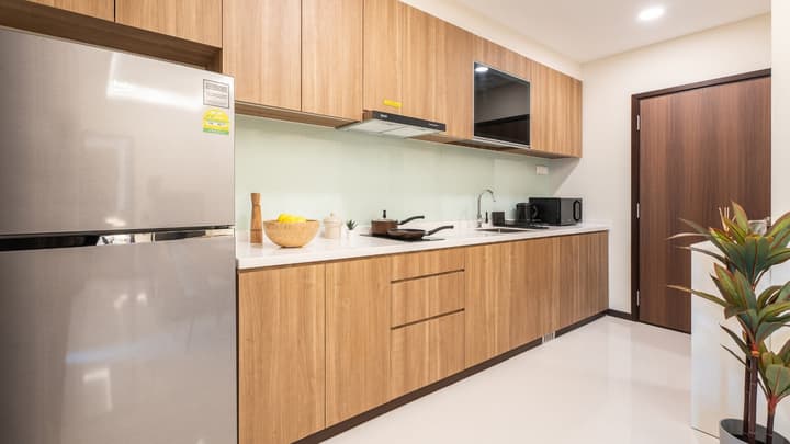 Modern kitchen with light wood-grain cabinets, white countertop, a stainless steel refrigerator, and a black cooktop and sink area.