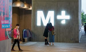 Exterior view of a building with a concrete facade featuring a large, brightly illuminated white M+ logo. Two people are standing beneath the sign, and a person wearing a pink backpack and face mask is walking past a poster for a Yayoi Kusama exhibition.