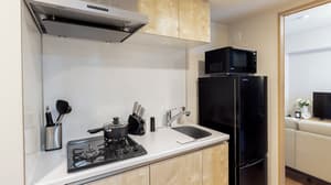 Compact kitchen area with light wood-effect cabinets, a white counter, a black gas cooktop, a stainless steel sink, a black refrigerator, and a microwave on top in Monzennakacho.