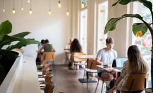 Blurred interior view of a bright cafe or communal lounge area, showing several patrons working or sitting at tables with large plants and hanging light fixtures.
