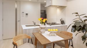 A dining area in a furnished apartment with a dark wood table set with flowers and fruit, providing a view into the compact kitchen with a cooktop and stainless steel hood.