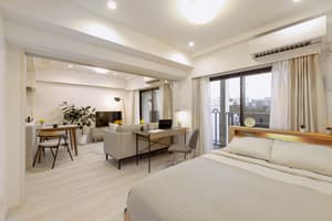 A wide-angle view of a bright, open-plan studio apartment showing the bedroom in the foreground, a dedicated work desk area, and a living/dining area visible through a wide doorway.