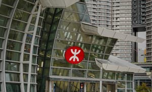 Exterior view of a modern building entrance constructed with white framework and angled glass panels. A large, iconic red and white circular MTR logo is centered on the facade, with tall residential buildings visible in the background.
