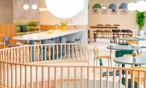 Wide view of a bright and spacious communal shared space. The space is furnished with a mix of long white bar-height tables, small round tables, and semi-circular booths with light wood slat railings.