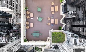 Aerial view of a rectangular, wood-decked rooftop terrace with various outdoor seating arrangements and green patches, surrounded by tall city buildings.