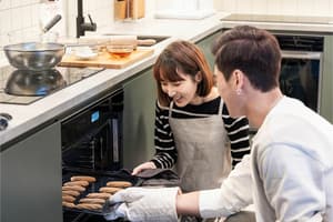 A smiling couple, a man and a woman, taking a tray of freshly baked cookies out of a modern oven in a shared coliving kitchen.