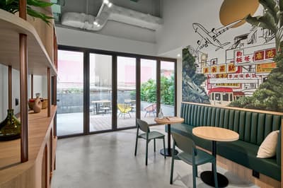 Bright communal living room with built-in green banquette seating, a Hong Kong-themed mural on the wall, and glass doors opening to an outdoor patio.