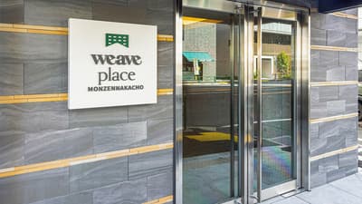 Exterior view of the main entrance to Weave Place Monzennakacho, featuring a contemporary grey slate wall with gold accents, a white sign with the green 'weave place' logo and name, and large glass double doors.