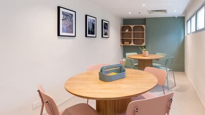 Bright communal workspace featuring two wooden round tables with pastel pink and mint-green chairs, set against a white and olive-green wall with framed photos.