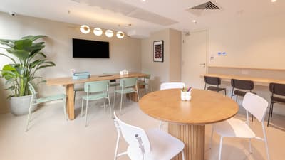 Multi-purpose room with a long wooden table for meetings or group work, a smaller round table, mint-green and white chairs, and a wall-mounted monitor.