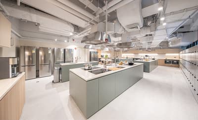 A spacious, commercial-style shared kitchen with multiple light green prep islands and stainless steel range hoods overhead. The room features several stainless steel refrigerators along one wall and a long wall of numbered storage lockers on the right. Exposed white ceiling ducts are visible above.