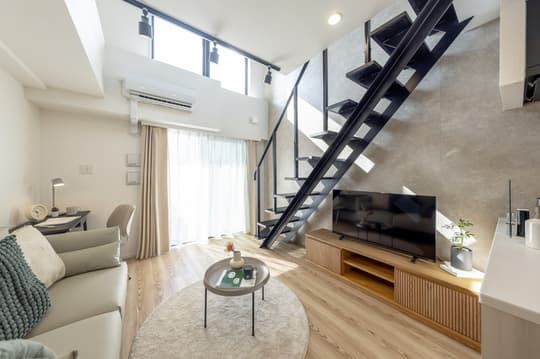 A bright, modern loft apartment living space featuring a comfortable sofa, a TV console, light wood floors, and an imposing black industrial staircase leading to the upper level.