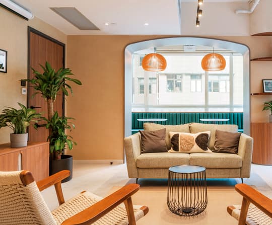 A cozy residential lounge area with a light gray sofa and wooden lounge chairs, centered around an arched window looking out to a street view with retro orange pendant lights.