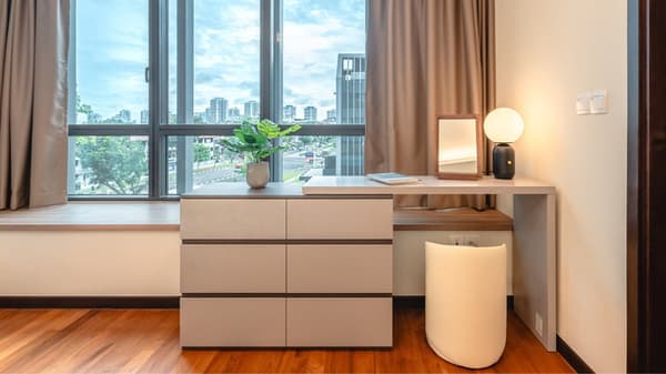 Bedroom area with a light grey dresser, integrated wooden desk and stool, positioned in front of a large window with a view of the city street.