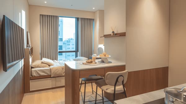 Bright, open-concept urban studio suite featuring a wooden kitchen bar/dining table with stools separating the cooking area from the cozy bedroom and large window.