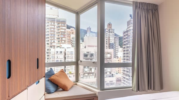 Interior view of a modern studio unit with a large corner window, built-in window seat/bench, and a tall, wood-grain storage cabinet on the left, offering a dense city view.