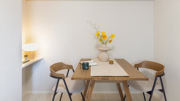 Close-up of the apartment's dining area featuring a wooden table with a vase of yellow flowers, two modern wooden chairs, and a small built-in shelf with a desk lamp in Monzennakacho.