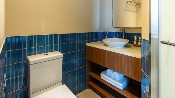 View of a compact, bright bathroom featuring a white toilet, a wall-mounted towel rack, and a sink vanity with a mirror above it, set against a wainscoting of blue mosaic tiles.