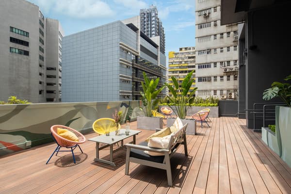 A sunny, modern rooftop terrace with wooden decking, a small lounge table, bright yellow and pink outdoor chairs, and large potted plants, set against a city backdrop.