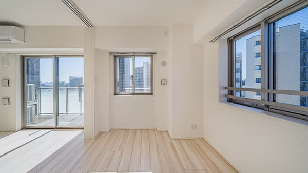 Sunlit, empty room with light wood-style flooring, two separate windows with safety screens, and a large sliding glass door leading to a balcony.