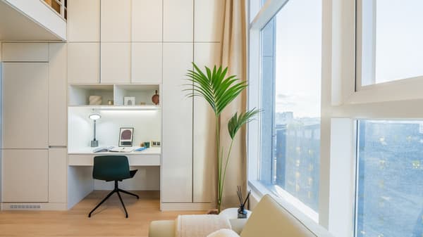 Close-up of a modern, built-in white desk and shelving unit with a dark green chair, next to a floor-to-ceiling window overlooking the city.
