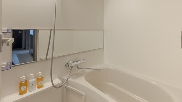 Clean white bathroom showing a deep bathtub, a shower attachment, a chrome faucet, a narrow mirror, and a shelf with toiletries in Monzennakacho.