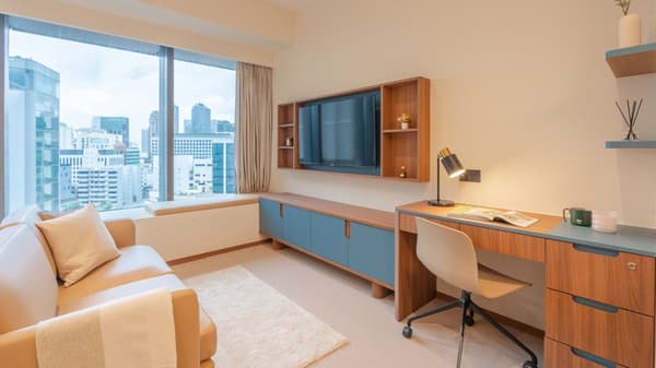 Light and modern studio apartment with a desk and chair, integrated blue and wood TV console, and large windows overlooking the city.
