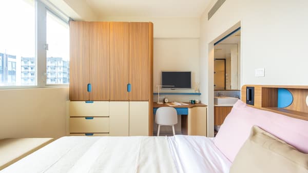 View over a queen bed toward a bright window wall with a large, wood-grain built-in wardrobe, a small desk with a chair and monitor, and a glimpse of the bathroom vanity.