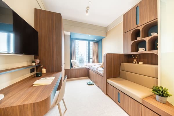 Interior view of a small room with multi-functional, built-in wood furniture. The space includes a desk, wall-mounted TV, cushioned seating with integrated storage, and a bed near a window with a city view.