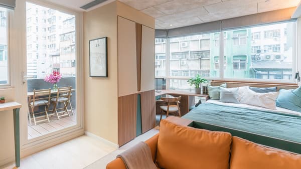 Modern studio apartment interior featuring a bed with teal and white bedding, an orange sofa in the foreground, a built-in desk, and a glass door leading to a small outdoor balcony with a table and chairs overlooking a dense city.