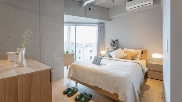 Bedroom with a light wood bed frame, white and mustard-colored bedding, and exposed concrete column and ceiling accents, with a large window and city view in the background.