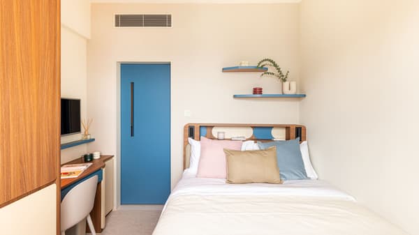 View of a compact studio featuring a queen-sized bed with wooden headboard, a pale blue door, a small desk/dressing area to the left, and floating blue accent shelves above the bed.