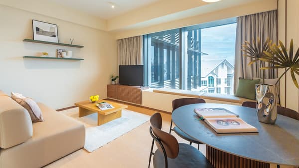 Open-concept living and dining area of the suite, showing a beige sofa, modern round dining table, and large window with a built-in window seat.