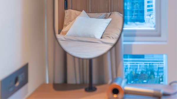 A close-up shot focused on a round, standing mirror on the bedroom desk, reflecting the bed and emphasizing the stylish wood finishes and large window light.
