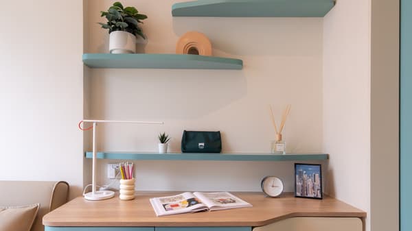 Close-up of a wooden desk and white wall with two floating mint-green shelves, decorated with a lamp, plants, books, and small decorative items.