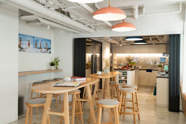 A shared dining area with light wooden high-top tables and stools, opening into a full, well-equipped kitchen visible through a foldable partition.