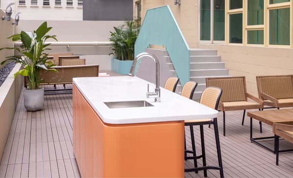 An outdoor patio area with wood decking, featuring a modern cooking island with a sink and an orange base, surrounded by black and rattan-style bar stools and outdoor furniture.