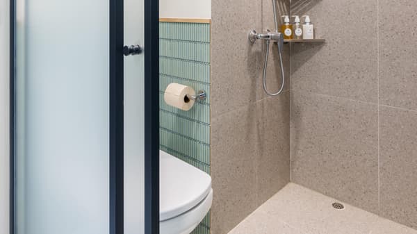 Compact bathroom with a clear partition separating the toilet area from the shower, featuring small turquoise ridged tile and large beige terrazzo-style shower tiles.