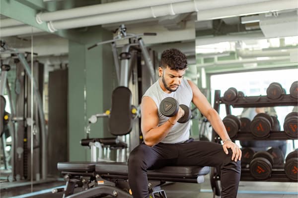 A focused young man sitting on a weight bench and performing bicep curls with a dumbbell in a brightly lit modern fitness center.