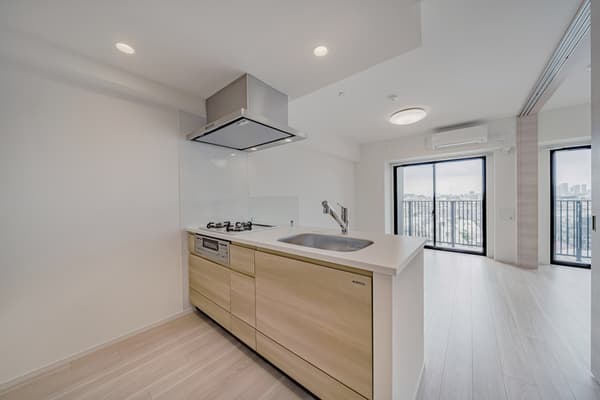 Modern kitchen with light wood cabinets, a white countertop, a stainless steel sink, a cooktop, and a silver range hood, looking toward the open living area and balcony.