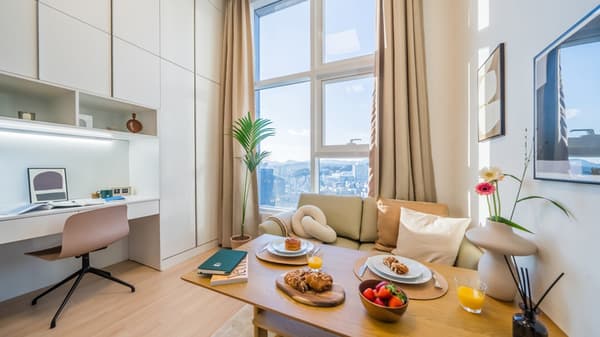 Sunlit apartment interior showing a combined living and dining area with a wooden table set for two, a built-in desk, and large windows with high ceilings.