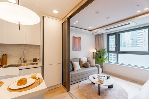 Modern, open-plan studio apartment showing a light gray sofa and round coffee table in the living area, separated from the compact white kitchen by a sliding glass door and view of a high-rise city through the window.