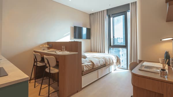 Modern studio apartment view showing the double bed with under-bed storage, a wall-mounted TV, a dividing counter with an integrated two-seater dining/bar area, and a large window with sheer curtains.